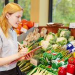 Junge Frau mit Klemmbrett und Stift in der Hand. Regal mit frischem Gemüse vor ihr. Als Frischespezialist/in übernimmst du Aufgaben im kaufmännischen und serviceorientierten Lebensmittelbereich. Beruf: Frischespezialist/in im Einzelhandel