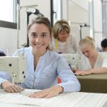 Eine junge Frau arbeitet an einer Nähmaschine. Im Hintergrund sitzen weitere junge Leute, ebenfalls an Nähmaschinen. Geschicklichkeit und ein gutes Augen sind für diesen Beruf von Vorteil. Beruf: Polster- und Dekorationsnäher/in