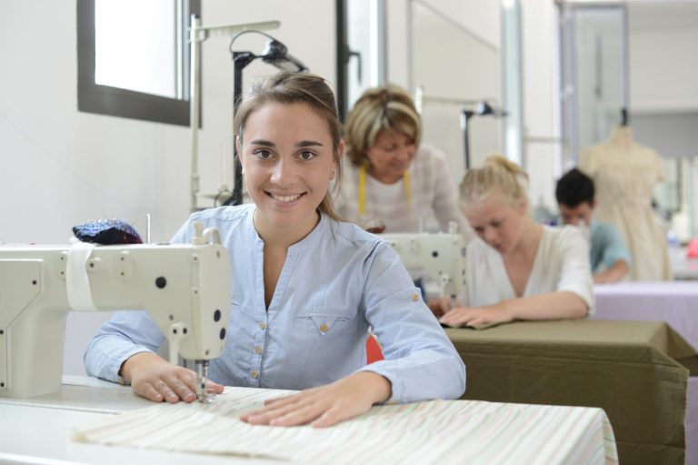 Eine junge Frau arbeitet an einer Nähmaschine. Im Hintergrund sitzen weitere junge Leute, ebenfalls an Nähmaschinen. Geschicklichkeit und ein gutes Augen sind für diesen Beruf von Vorteil. Beruf: Polster- und Dekorationsnäher/in