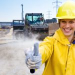 Eine junge, freundliche Frau in Schutzbekleidung steht vor einer Straßenbaustelle. Mit der rechten Hand zeigt sie den Daumen hoch. Die Ausbildung zum Straßenbauer bietet langfristige Perspektiven. Beruf: Straßenbauer/in